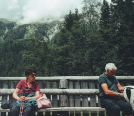 Two people sitting on a long wooden bench in a mountainous forest setting, with one person holding hiking poles and the other looking into a backpack