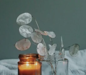 two jars next to each other, one with a lit candle, and one with white silk flower leaves on stems