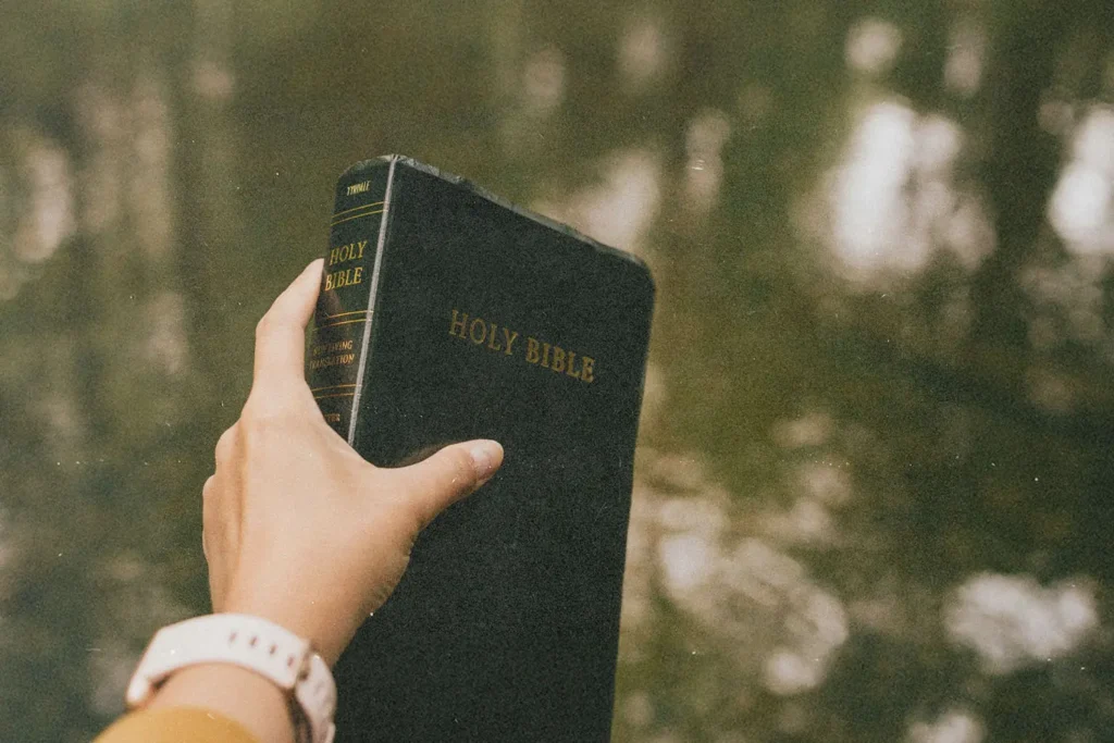 A hand holds up a black Holy Bible outdoors, with calm water reflecting soft light in the background