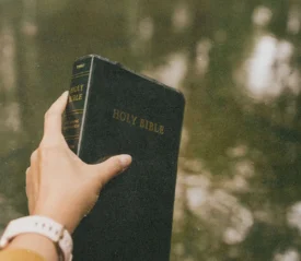 A hand holds up a black Holy Bible outdoors, with calm water reflecting soft light in the background