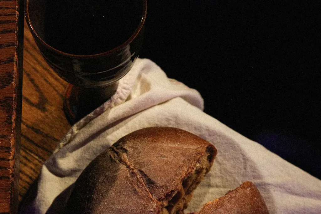 broken bread on a cloth napkin next to a communion cup on the edge of a wooden table