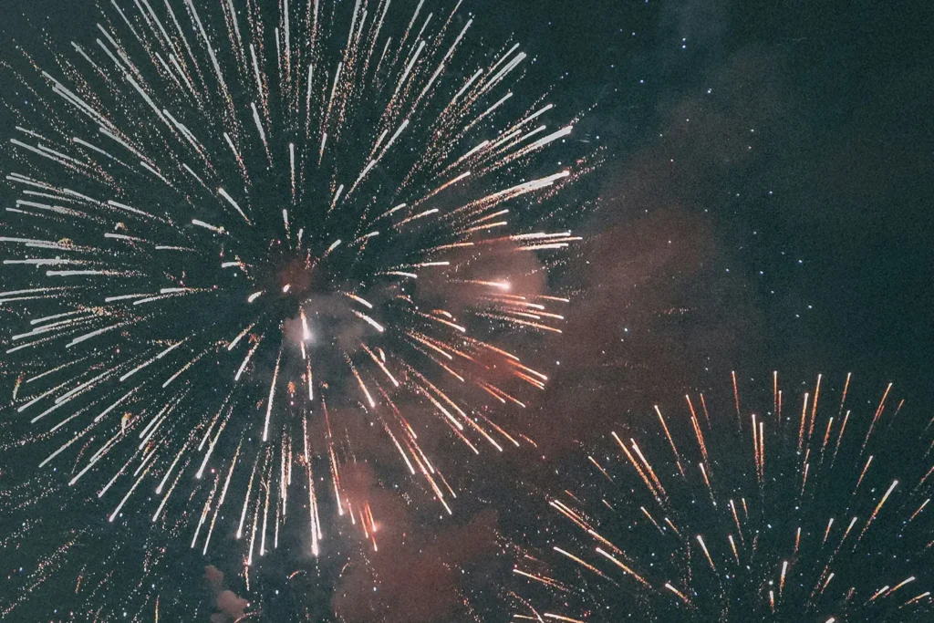 A night sky filled with large bursts of fireworks in bright white and orange tones