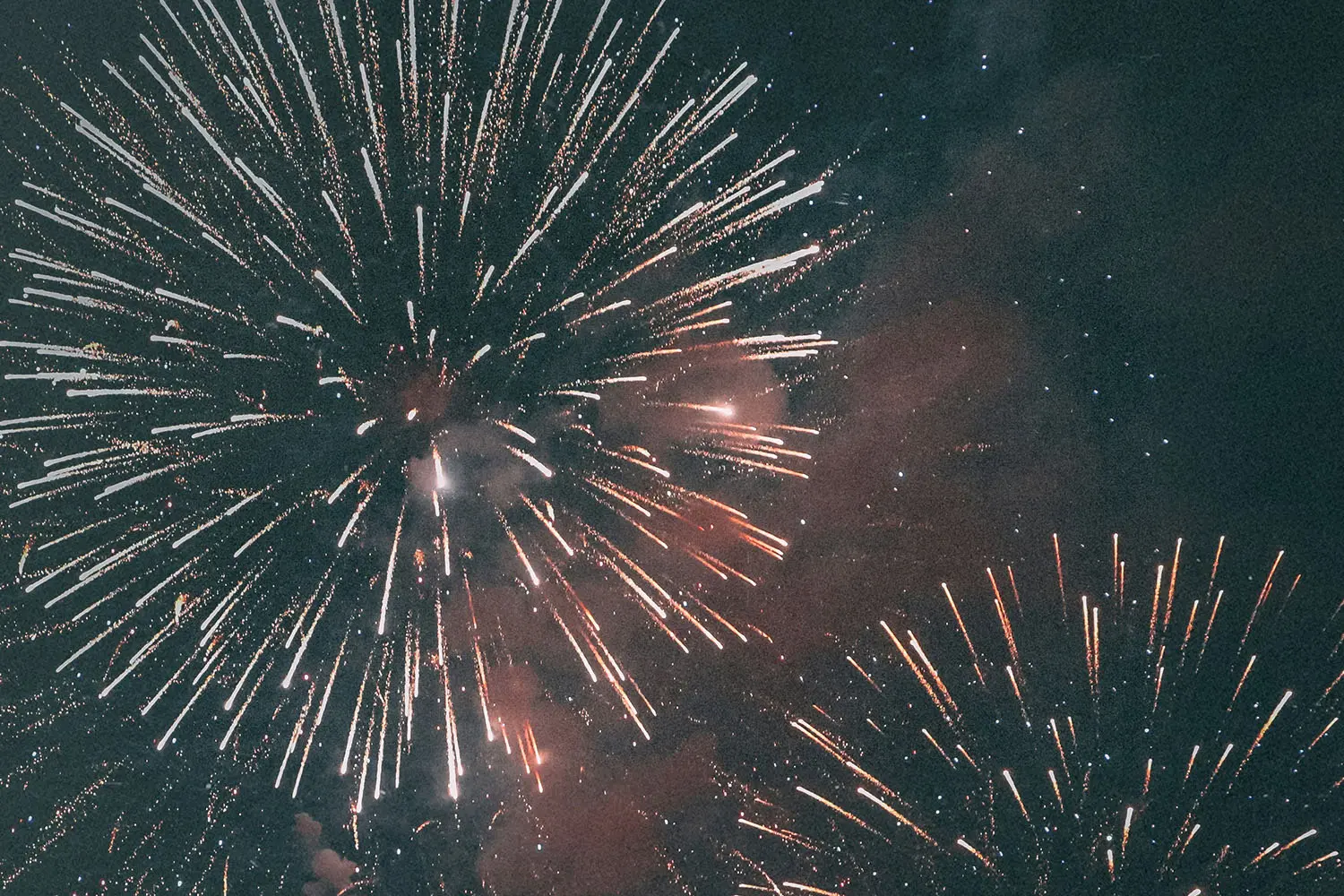 A night sky filled with large bursts of fireworks in bright white and orange tones