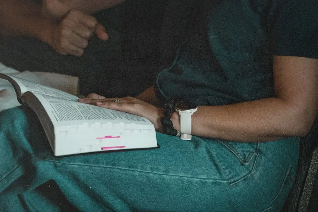 A person sits with an open book resting on their lap, their hand placed on the pages, wearing a watch and denim jeans