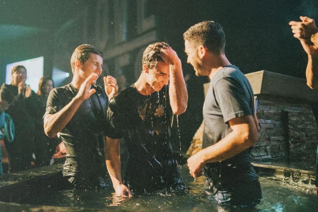 A group of people stand in a shallow pool during a baptism ceremony, water dripping as one person touches their forehead
