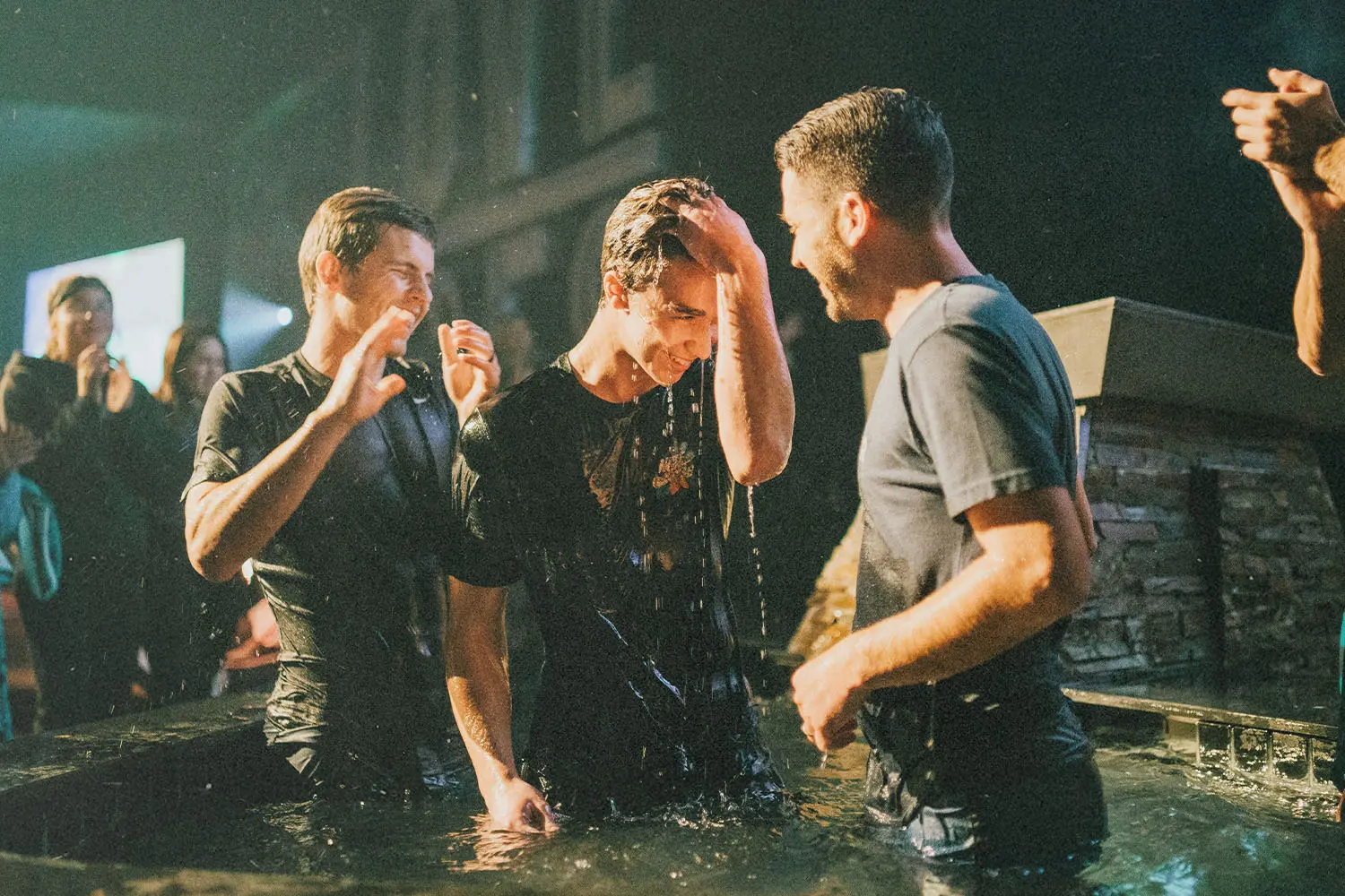 A group of people stand in a shallow pool during a baptism ceremony, water dripping as one person touches their forehead