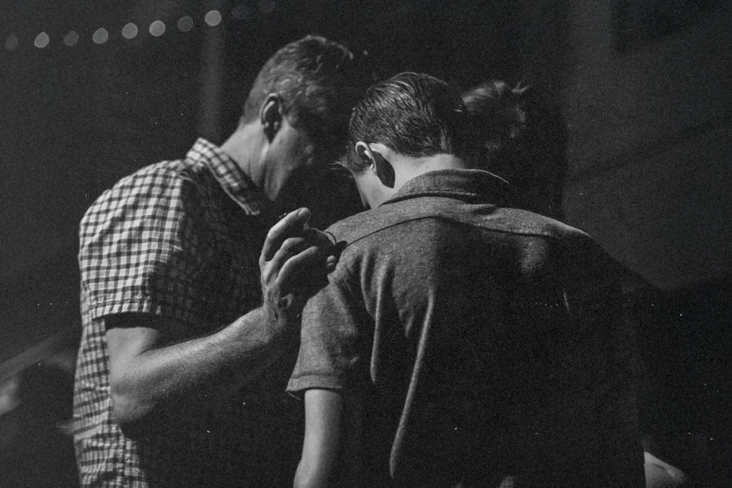 Three people stand closely together in dim lighting, with one person placing a hand gently on another’s shoulder in a moment of support or prayer