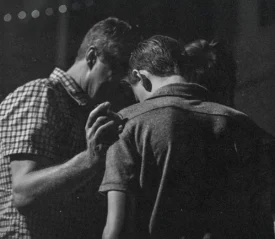 Three people stand closely together in dim lighting, with one person placing a hand gently on another’s shoulder in a moment of support or prayer