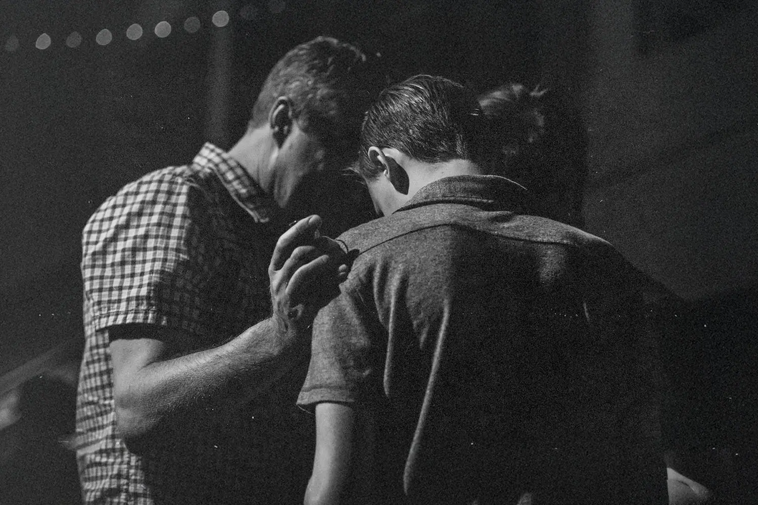 Three people stand closely together in dim lighting, with one person placing a hand gently on another’s shoulder in a moment of support or prayer