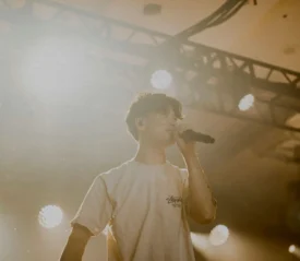 A performer holds a microphone on a brightly lit stage, surrounded by warm spotlights and overhead rigging