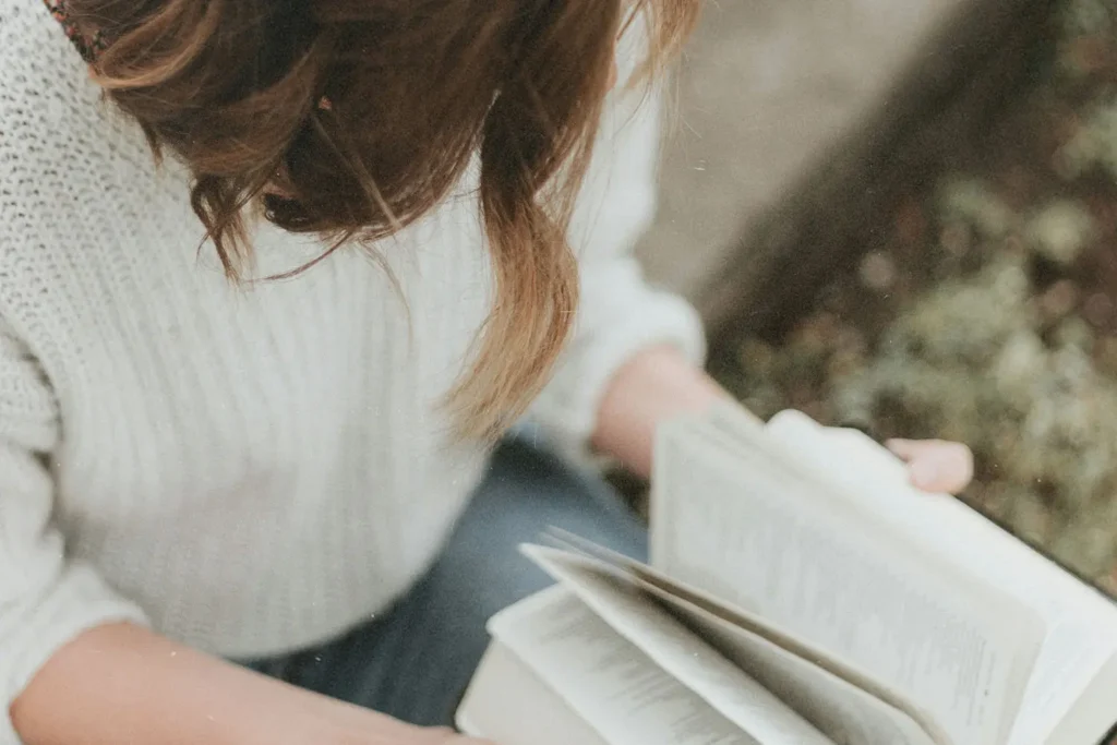A person in a white sweater reads a book outdoors, holding the pages open across their lap