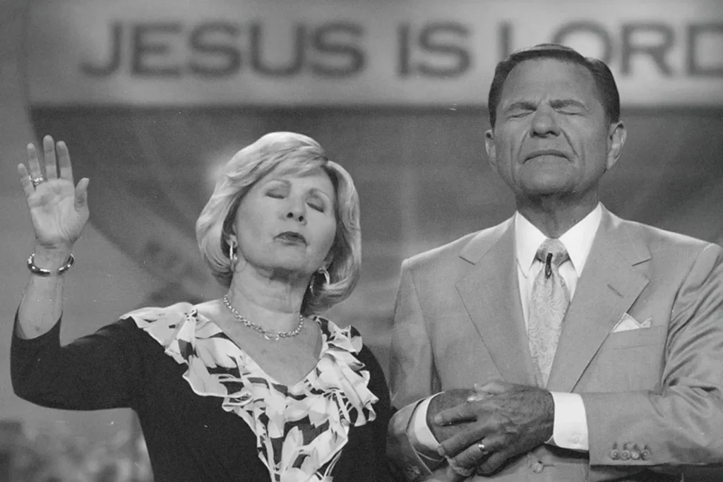 Kenneth and Gloria Copeland stand together in front of a backdrop that reads “JESUS IS LORD,” raising hands in a gesture blessing