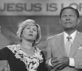 Kenneth and Gloria Copeland stand together in front of a backdrop that reads “JESUS IS LORD,” raising hands in a gesture blessing