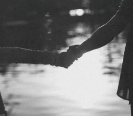 Two people hold hands beside a reflective body of water, shown as a soft black‑and‑white silhouette