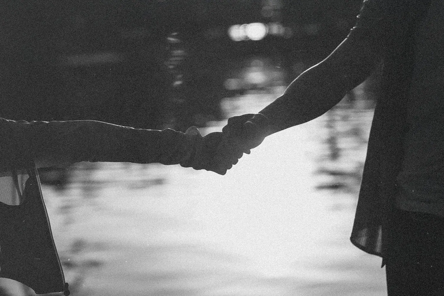 Two people hold hands beside a reflective body of water, shown as a soft black‑and‑white silhouette