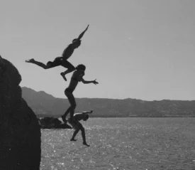 Three people leap from a rocky cliff into the water below, captured mid‑air in a black‑and‑white scene
