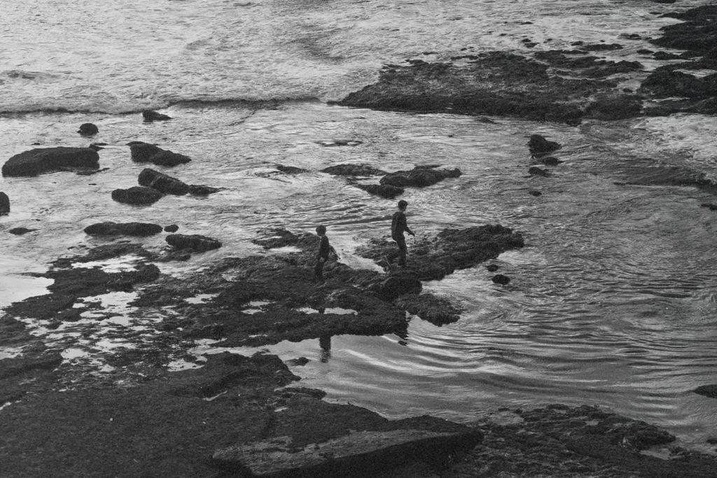 Two children walk along rocky tide pools at the edge of the ocean as waves roll in