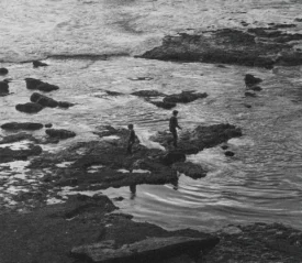 Two children walk along rocky tide pools at the edge of the ocean as waves roll in