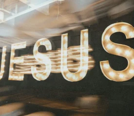 Large illuminated letters spelling “JESUS” glow with marquee-style bulbs against a dark background, with motion blur trailing from the lights
