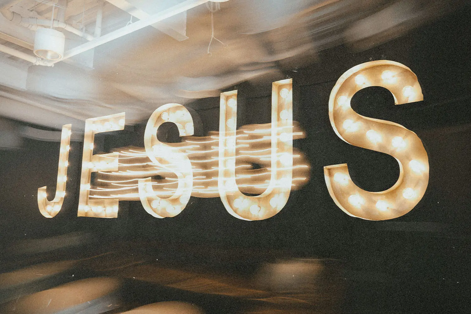 Large illuminated letters spelling “JESUS” glow with marquee-style bulbs against a dark background, with motion blur trailing from the lights