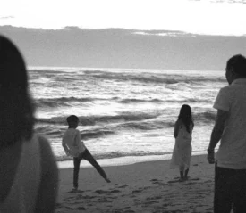 A black-and-white scene of a family standing on a beach at dusk, watching waves roll onto the shore