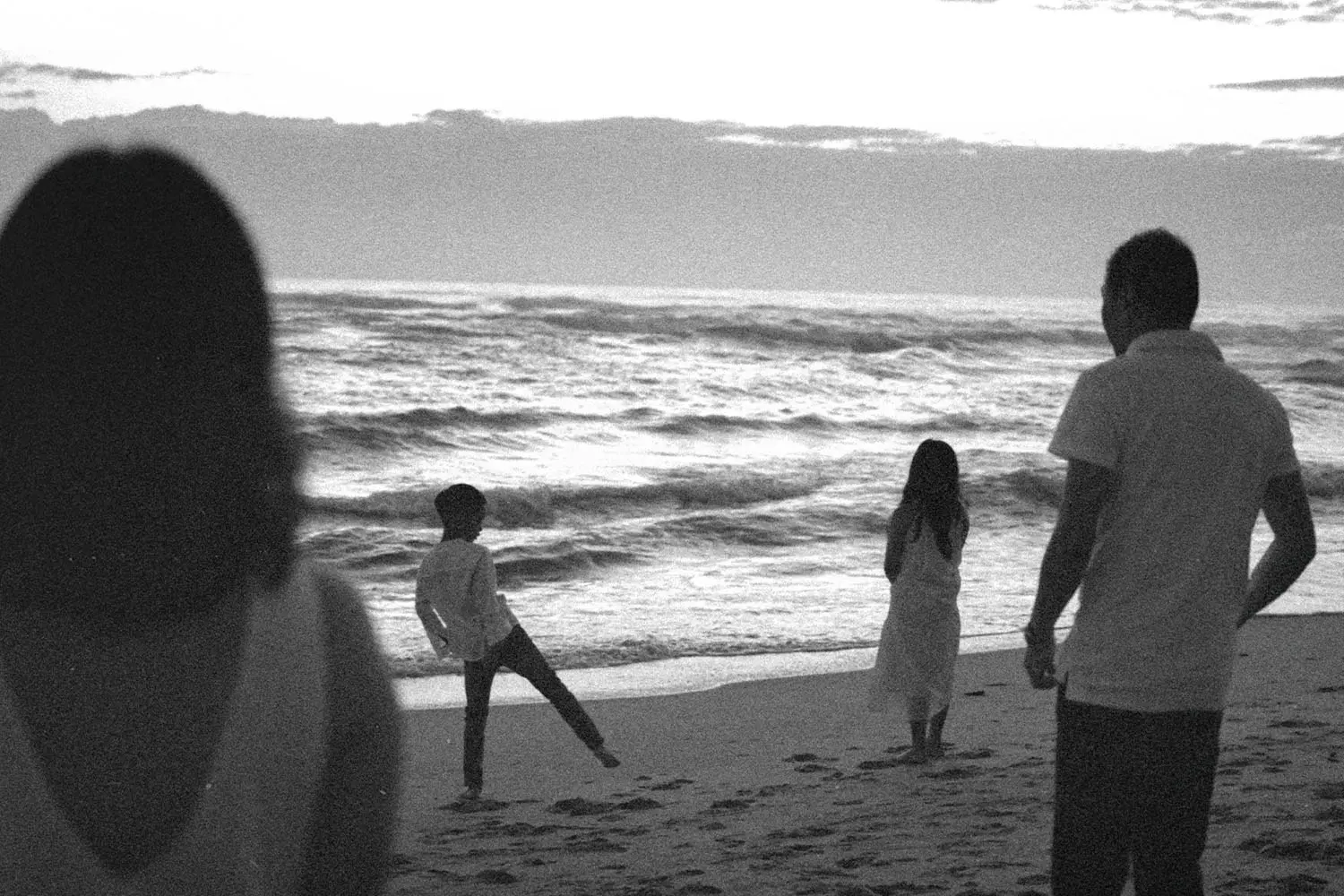 A black-and-white scene of a family standing on a beach at dusk, watching waves roll onto the shore