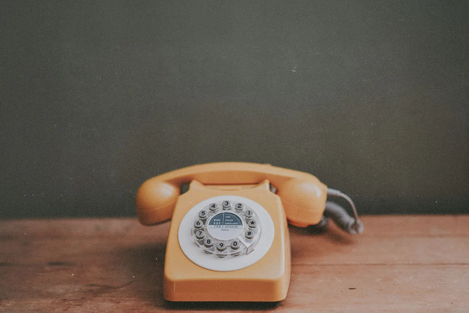 A vintage yellow rotary telephone sits on a wooden surface in front of a muted green wall