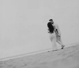 A couple stands close together on a quiet beach, holding hands as they face each other beside the shoreline