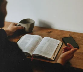 Man reading open Bible resting on wooden table with a coffee cup in hand