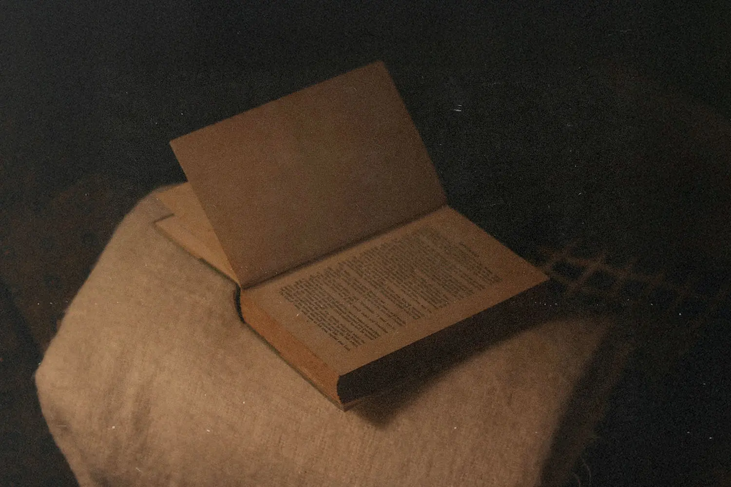 An old, open book rests on a textured cloth surface in dim, warm lighting