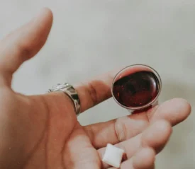 A hand holds a small cup of red communion juice with a white communion wafer resting near the fingertips