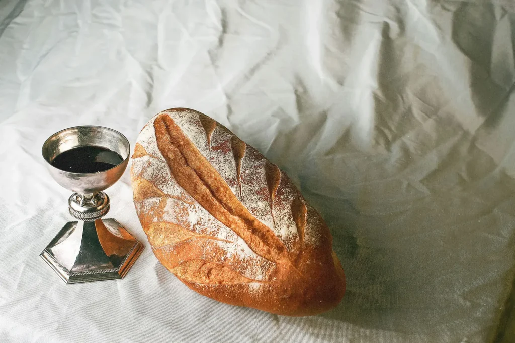 Loaf of rustic bread sits beside a silver communion cup filled with dark liquid on a softly wrinkled white cloth