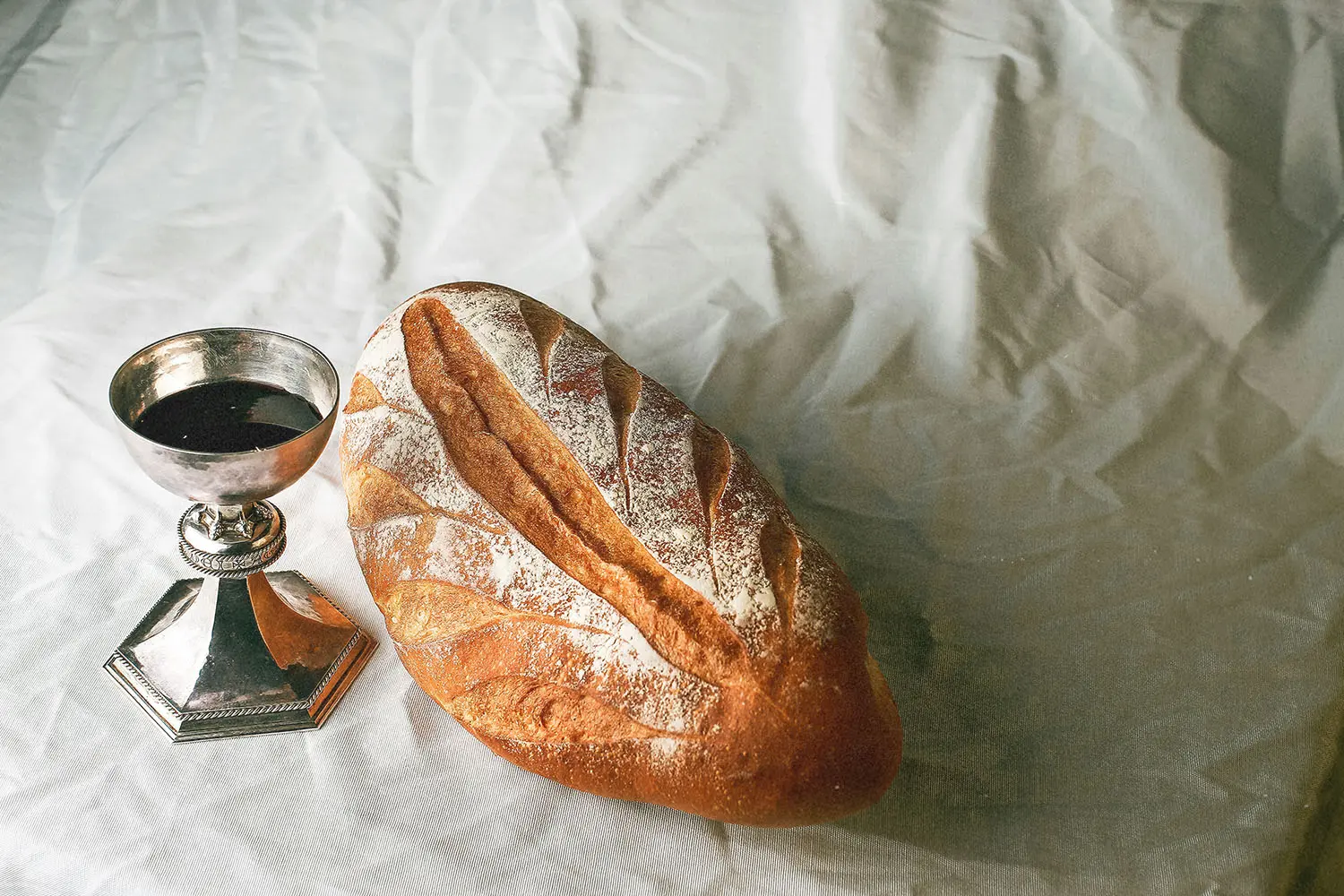Loaf of rustic bread sits beside a silver communion cup filled with dark liquid on a softly wrinkled white cloth