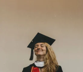 A graduate wearing a cap and gown stands against a plain beige background