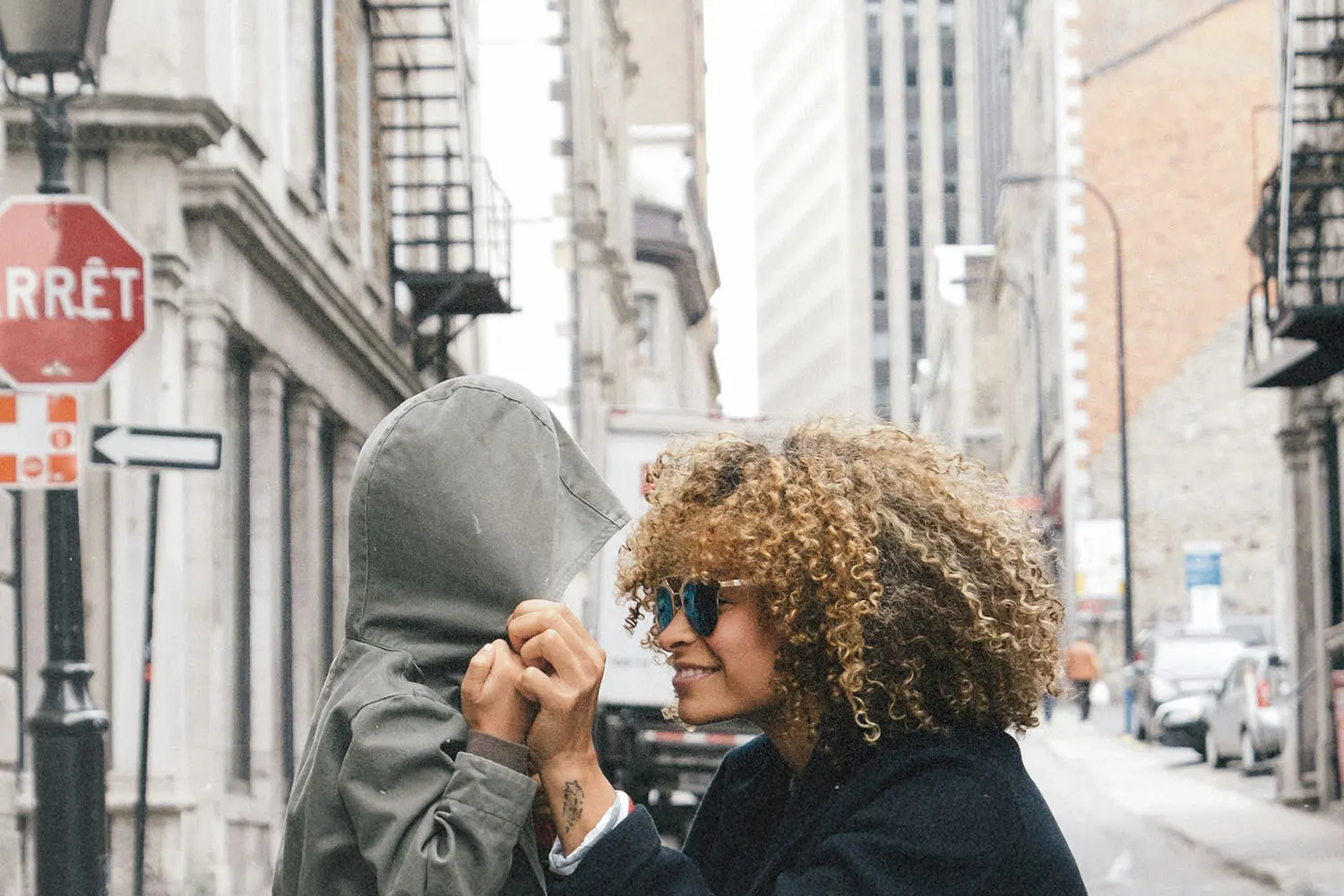 A person adjusts the hood of a child’s jacket on a city street lined with tall buildings, with a red French “Arrêt” stop sign visible in the background