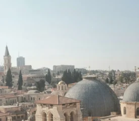 Cityscape featuring domed church roofs, stone buildings, and distant towers under a bright, clear sky