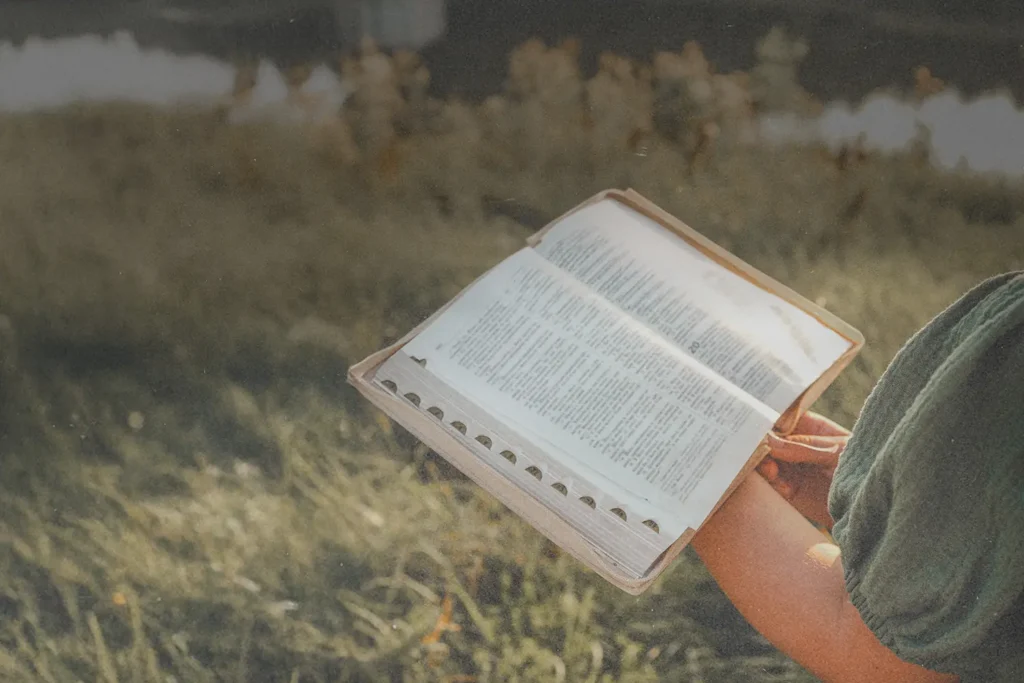 Open Bible held outdoors above sunlit grass near a calm body of water