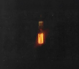 Single glowing Edison-style light bulb suspended in darkness, casting a warm orange filament glow