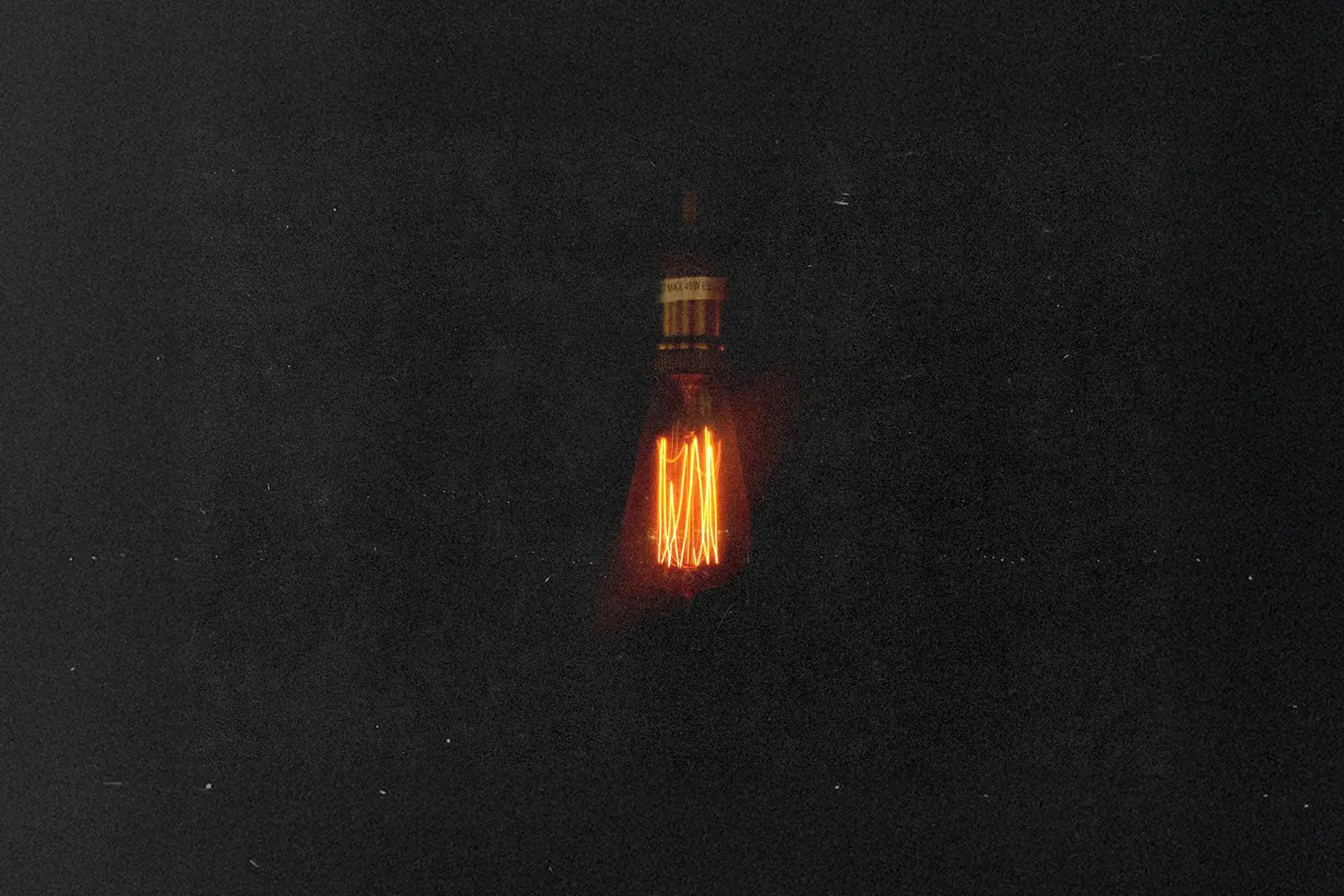 Single glowing Edison-style light bulb suspended in darkness, casting a warm orange filament glow