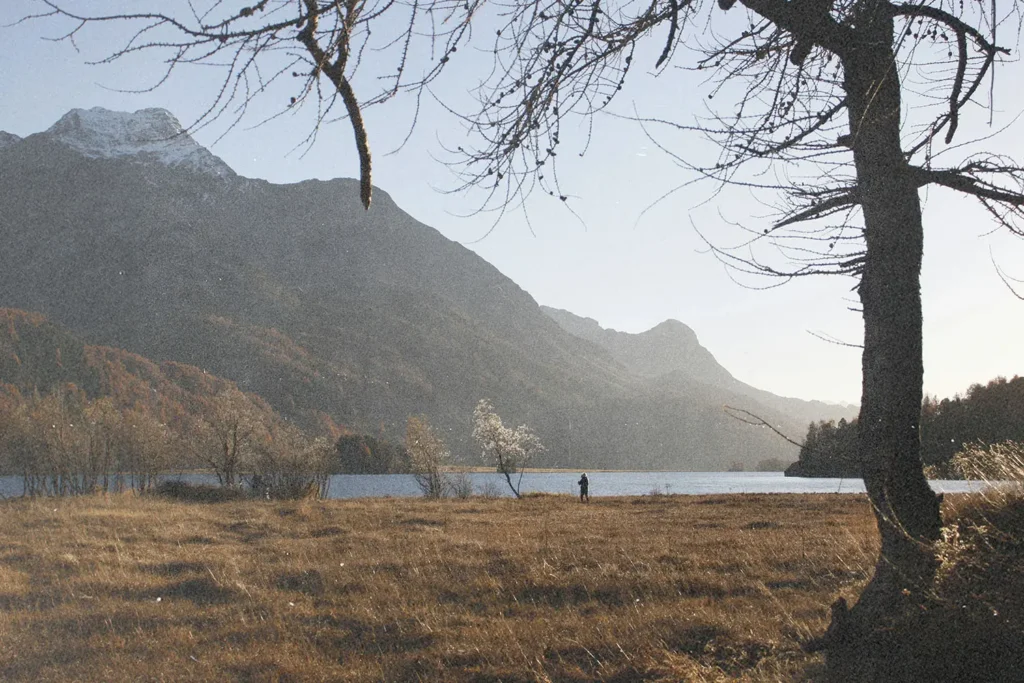 Wide landscape of a grassy field beside a lake with tall mountains in the background and a person standing near the shoreline