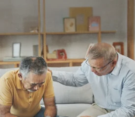 Person sitting on a couch with another person beside them offering support with a hand on their shoulder in a living‑room setting