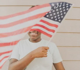 Young kid standing against a light-colored wall, waving an American flag that fills the foreground