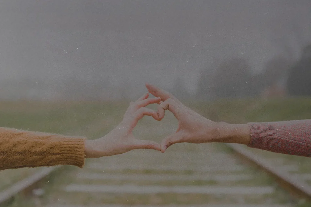 Two people extending their arms to form a heart shape with their hands over blurred railroad tracks