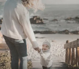 Adult holding a small child’s hand while walking down a wooden path toward a rocky beach with waves in the background