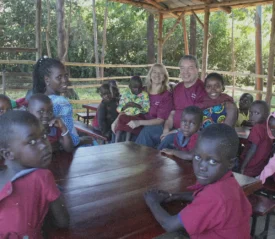 Children gather around missionaries seated at outdoor tables during a Uganda missions class, reflecting their commitment to teaching and discipleship.