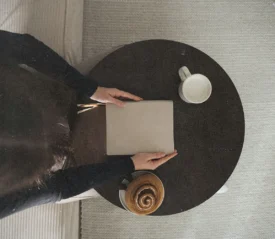 A person sits at a small round table with a closed notebook, a mug of coffee, and a pastry in warm natural light