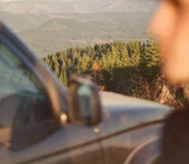 Parked car is shown in the foreground while a scenic landscape of rolling hills and evergreen forests stretches into the distance
