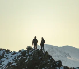 Two people stand on a snowy mountain ridge at sunrise or sunset, silhouetted against a pale sky