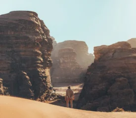 Person standing on a sandy desert path surrounded by towering rock formations under a bright sky