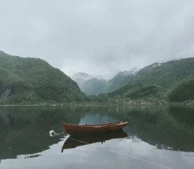 Wooden rowboat floating on a still lake surrounded by steep, forested mountains beneath an overcast sky